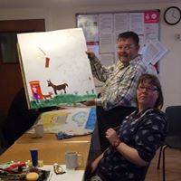 Adrian standing up holding a half-completed painting, with Kelly sat in front.